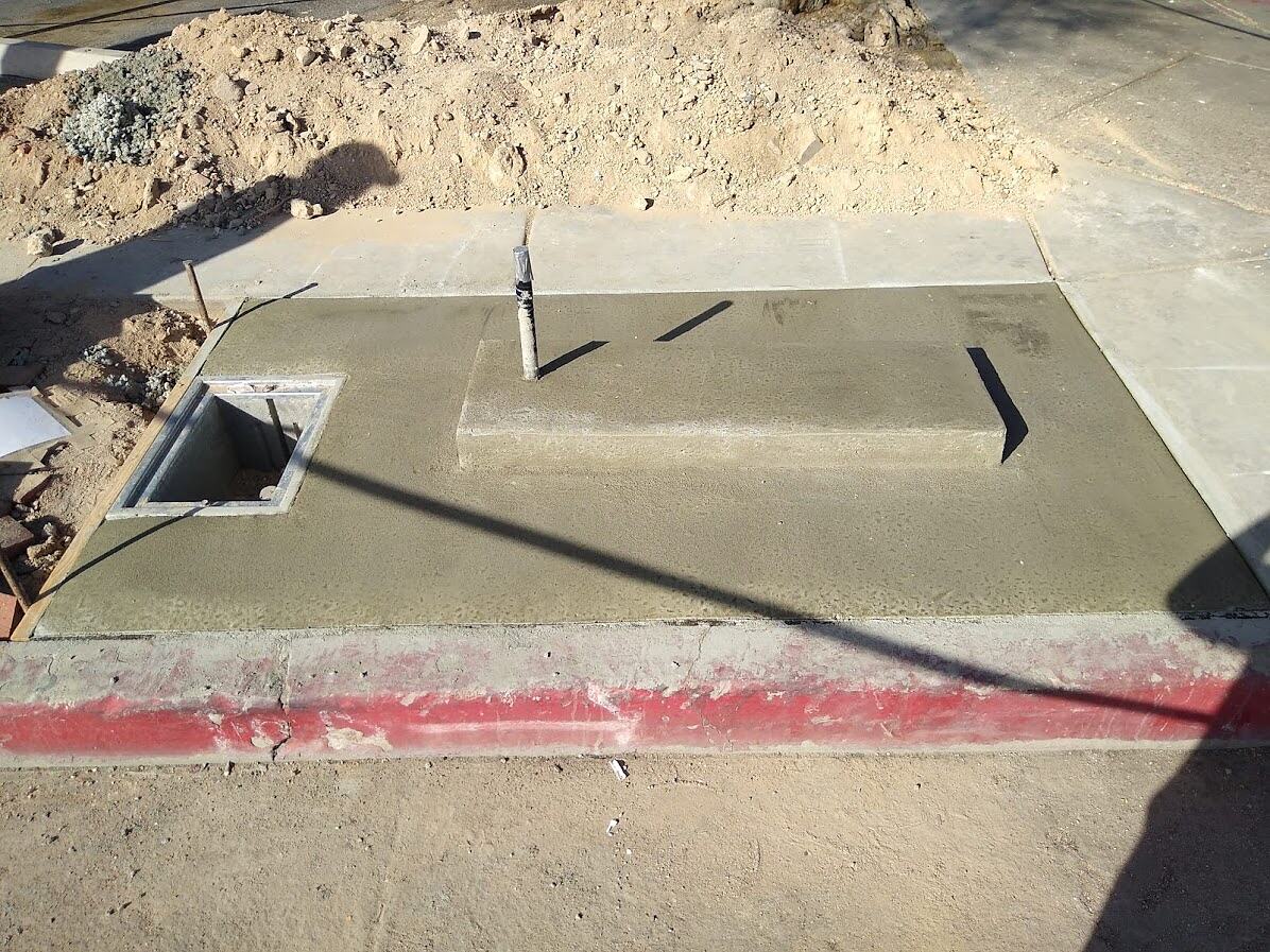 Finished concrete kiosk foundation with raised plinth, conduit stub-up and open pull box in a restored sidewalk